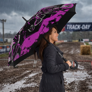 A woman in a dark raincoat stands in the rain at a muddy motocross track holding an open umbrella with a black background and a pink, black, and white graphic pattern.