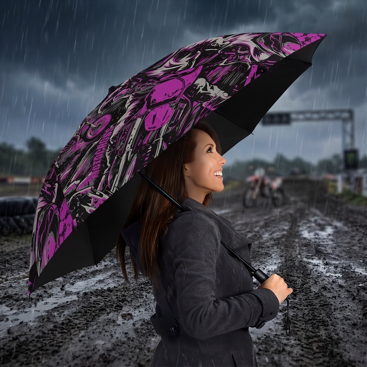 A woman in a dark gray coat stands in the rain holding a large open umbrella with a bold black, white, and bright pink graphic print. The background shows a blurred rainy outdoor scene with a dirt road and overcast sky.