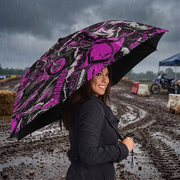 A woman in a dark coat stands in the rain holding a large open umbrella with a bold pink, black, and white graphic pattern. The background shows a muddy outdoor event or track with banners and blurred figures.