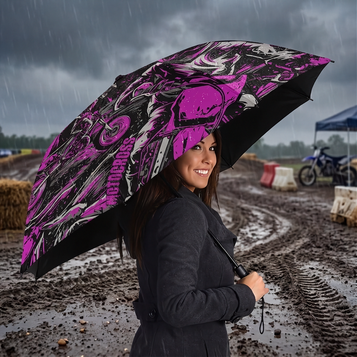 A woman in a dark coat stands in the rain holding a large open umbrella with a bold pink, black, and white graphic pattern. The background shows a muddy outdoor event or track with banners and blurred figures.