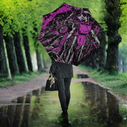 A person walking in the rain along a tree-lined path, holding a large open umbrella with a bold black and magenta graphic print. The scene is viewed from behind, with raindrops falling and reflections visible in the puddle.