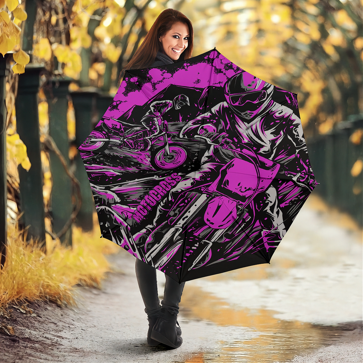 A woman holding an open umbrella in an outdoor autumn setting. The umbrella features a bold graphic print of a motorcyclist in pink, black, and white, with the word 'Motocross' printed vertically on the side.