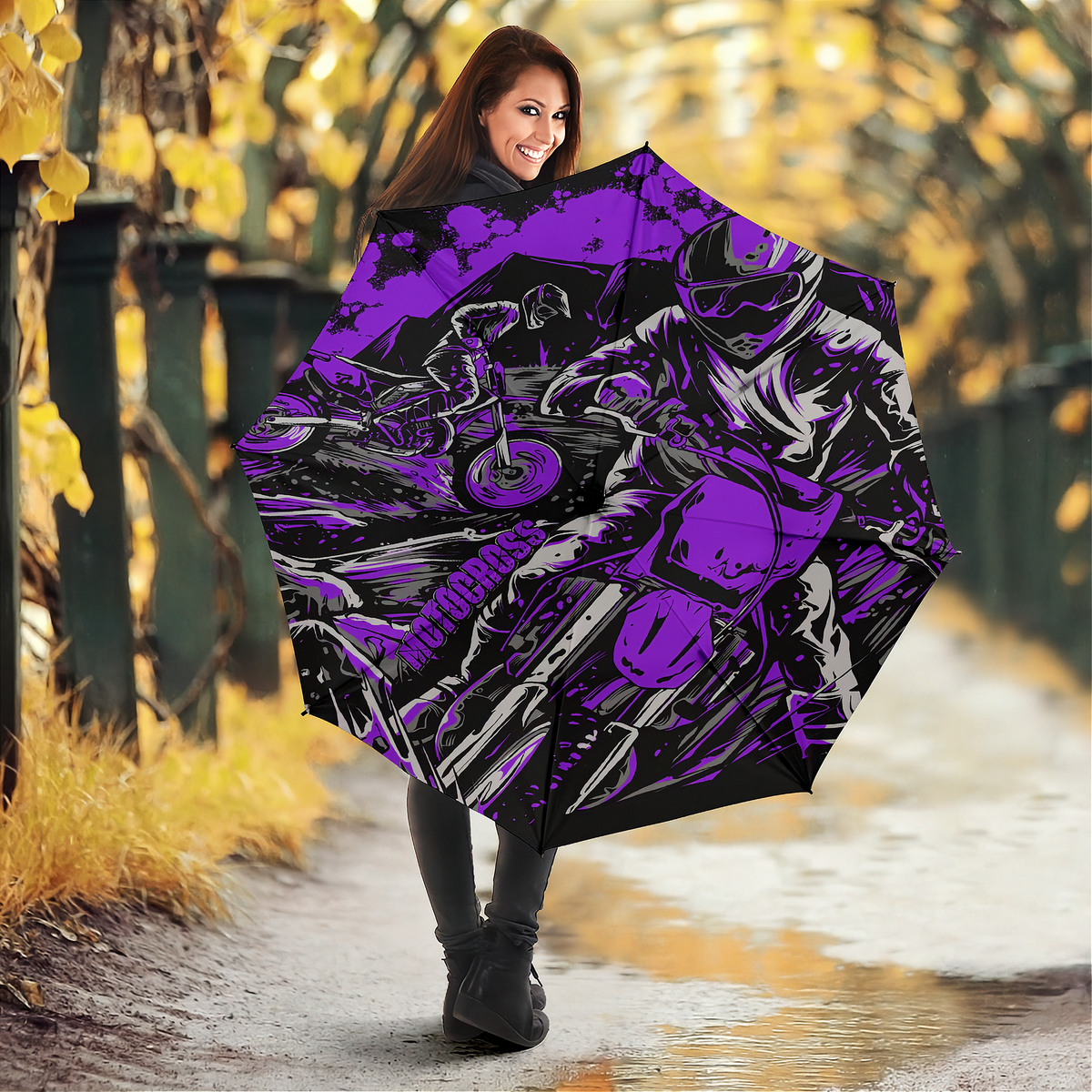 A woman standing on a wet path in an autumn park holding a large open umbrella. The umbrella features a bold graphic print of a motorcyclist in purple, black, and white.