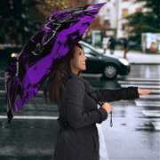 A woman in a dark gray coat stands in the rain holding a large open umbrella with a bold purple and black graphic design. The background shows a blurred city street with a car and pedestrians.