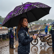 A woman in a black motorcycle jacket stands under a black and purple patterned umbrella on a muddy racetrack, with a dirt bike and other riders in the background.