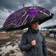 A man in a dark rain jacket stands in the rain holding a large open umbrella with a bold purple, black, and white graphic pattern. The background shows a muddy track under a dark, rainy sky.