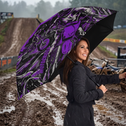 A woman in a black raincoat stands in the rain on a muddy dirt track, holding a large open umbrella with a bold purple, black, and gray graphic print featuring a skull and motorcycle design.