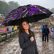 A woman in a gray coat holding a black umbrella with a purple and black graphic pattern, standing in the rain on a muddy race track with sponsor banners in the background.