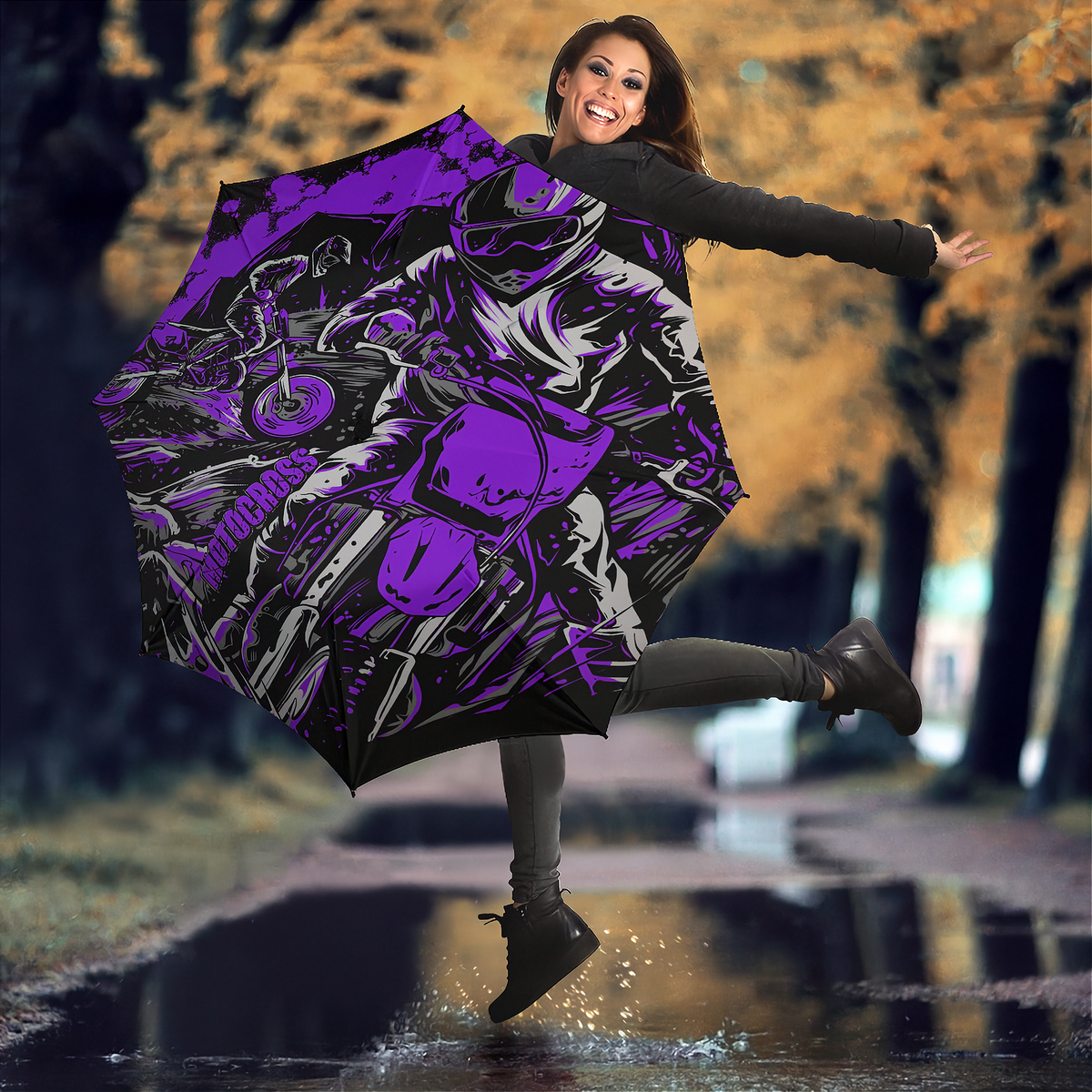 A woman jumps in a puddle outdoors holding a large open umbrella with a bold purple, black, and gray motorcycle graphic. The background shows blurred trees with autumn-colored leaves.
