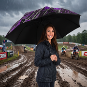 A woman smiling under a large black umbrella with a purple and white graphic pattern, standing in the rain on a muddy dirt track with motocross riders in the background.