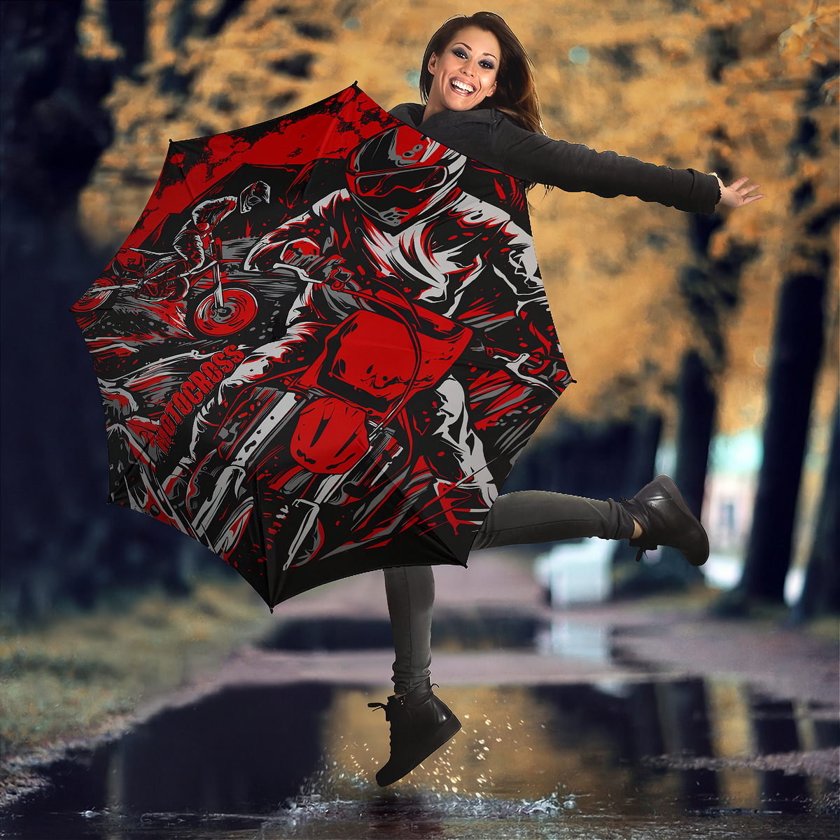 A woman jumps in a puddle outdoors while holding open a large umbrella printed with a bold red, black, and gray motorcycle graphic. Trees with yellow autumn foliage are blurred in the background.