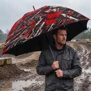 A man in a dark raincoat stands in the rain holding a large black umbrella with a bold red and white graphic print, set against a muddy outdoor track background.
