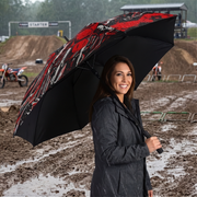 A woman in a dark raincoat stands in the rain at a muddy motocross track holding an open umbrella with a black background and a red, black, and white graphic pattern.