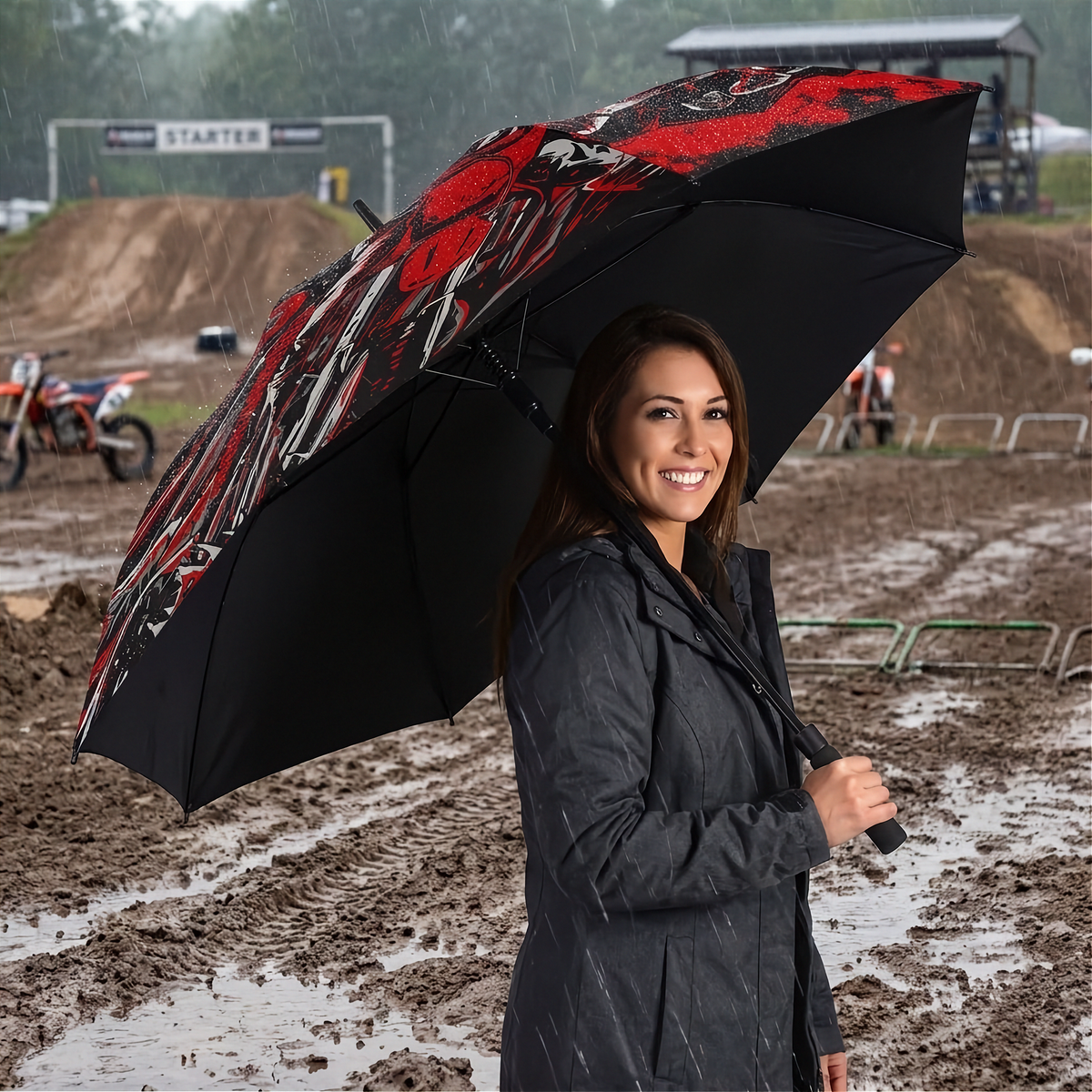 A woman in a dark raincoat stands in the rain at a muddy motocross track holding an open umbrella with a black background and a red, black, and white graphic pattern.