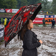 A woman in a dark coat stands in the rain holding a large open umbrella with a bold red, black, and white graphic pattern. The background shows a muddy outdoor event or track with banners and blurred figures.