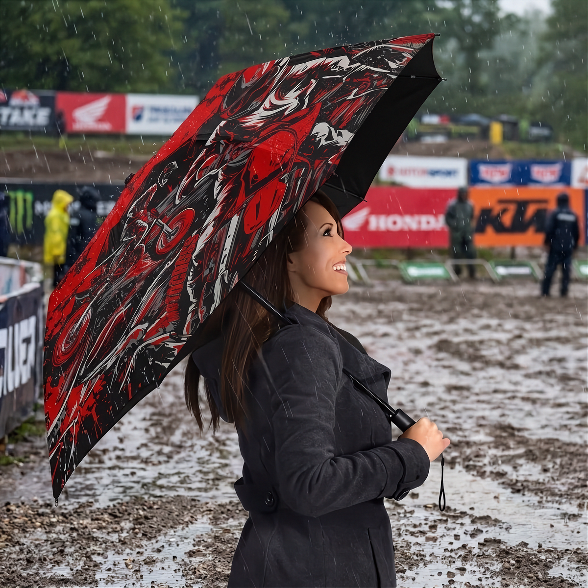 A woman in a dark coat stands in the rain holding a large open umbrella with a bold red, black, and white graphic pattern. The background shows a muddy outdoor event or track with banners and blurred figures.