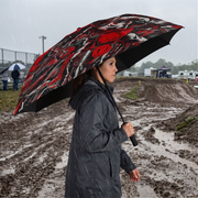 A woman in a dark gray raincoat stands in the rain holding a large open umbrella with a bold red, black, and white graphic pattern. The background shows a muddy outdoor path and a cloudy sky.