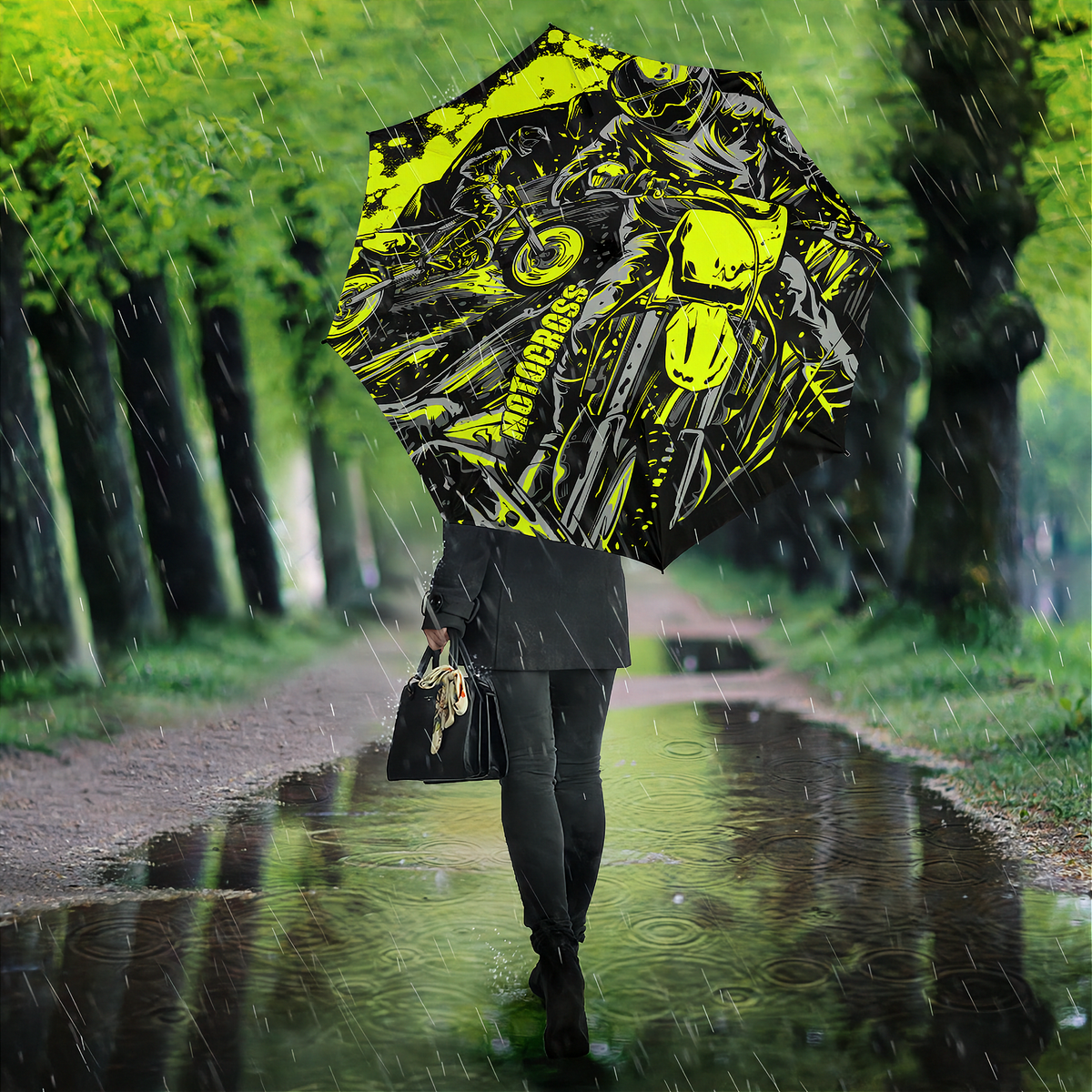 A person walking in the rain on a tree-lined path holding a large black and neon yellow graphic umbrella. The umbrella features a bold motorcycle-themed print with the word 'MOTOCROSS' visible.