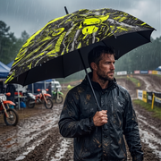 A man in a black rain jacket stands in the rain holding a large black umbrella with a bright yellow graphic print. The background shows a muddy outdoor motocross track with dirt bikes and a cloudy sky.