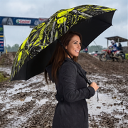 A woman in a dark gray coat holds a black umbrella with a bright yellow and black motocross graphic pattern while standing in a muddy outdoor track during rain. Castrol and other track signage are visible in the background.