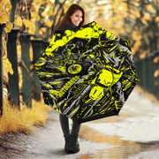 A woman standing on a wet path in an autumn park holding a large open umbrella. The umbrella features a high-contrast graphic print of a motorcyclist in neon yellow, black, and gray.