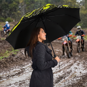 A woman in a dark raincoat holds a large black umbrella with a bright yellow graphic pattern while riding in the rain. Dirt bikes and riders are visible in the muddy background.