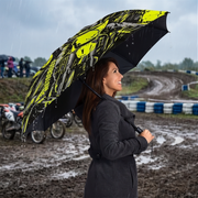 A woman in a dark gray coat stands in the rain at a muddy outdoor track holding a large black umbrella with a bright yellow and gray graphic print.