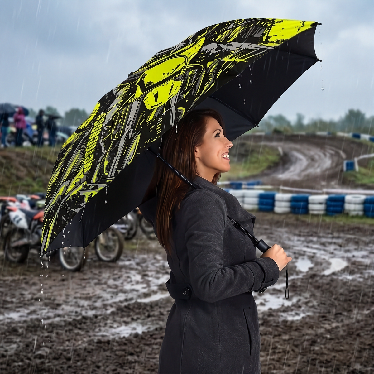 A woman in a dark gray coat stands in the rain at a muddy outdoor track holding a large black umbrella with a bright yellow and gray graphic print.