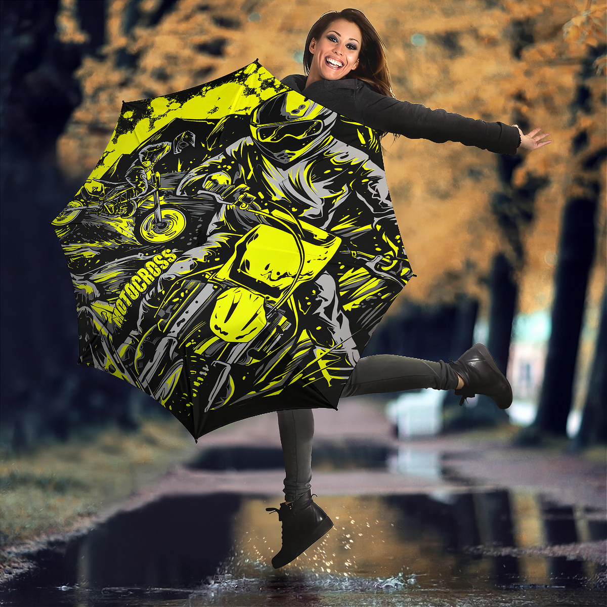 A woman jumps in a puddle outdoors while holding open a large umbrella printed with a bold black, gray, and neon yellow motorcycle design. Trees with autumn-colored leaves are blurred in the background.