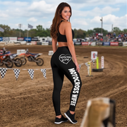 A woman stands on a dirt motocross track wearing black leggings with white 'MOTOCROSS WIFE' text and a heart logo on the back, along with a black sports bra. Dirt bikes and a blurred racetrack are visible in the background.