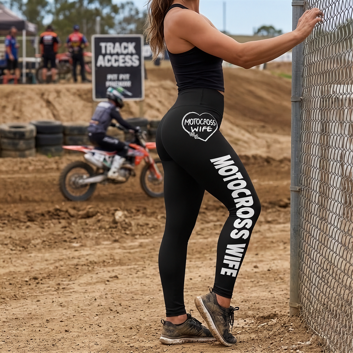 A woman stands beside a chain-link fence at a dirt motocross track, wearing black leggings with large white 'MOTOCROSS WIFE' text and a heart-shaped 'MOTOCROSS WIFE' logo on the upper thigh. A motocross rider is visible in the background.