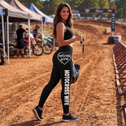 A woman stands on a dirt motocross track wearing black leggings with white 'MOTOCROSS WIFE' text and a heart graphic on the hip, along with a matching black sports bra. Tents, bikes, and track barriers are visible in the background.