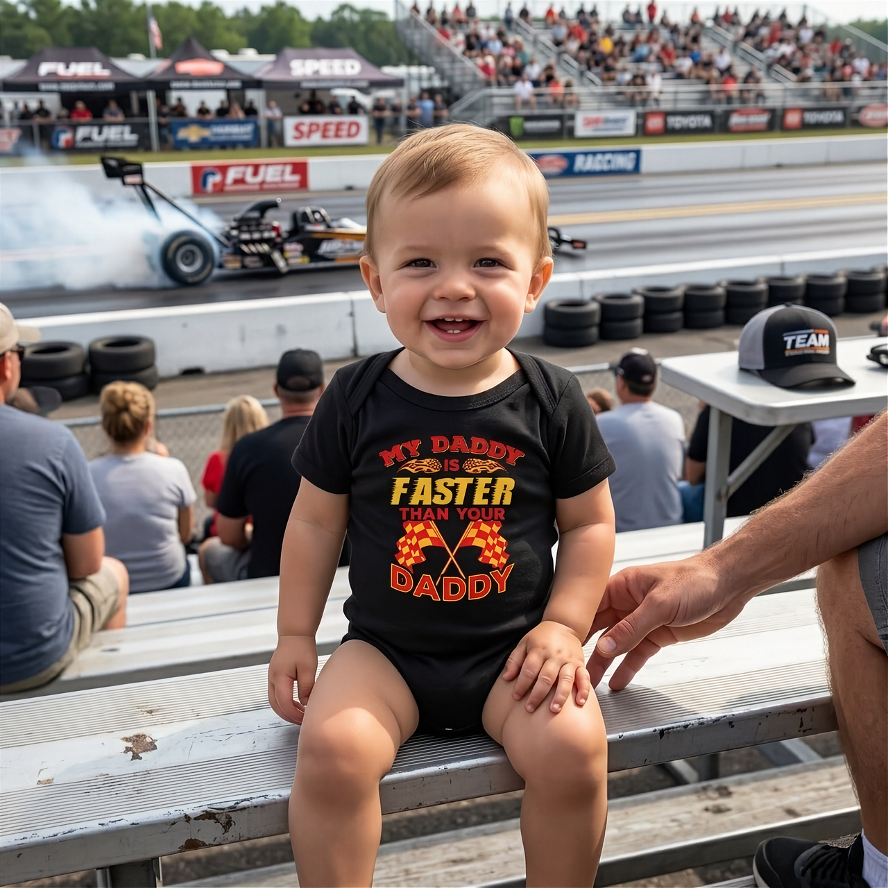 Child wearing a black onesie with text at a drag race track event