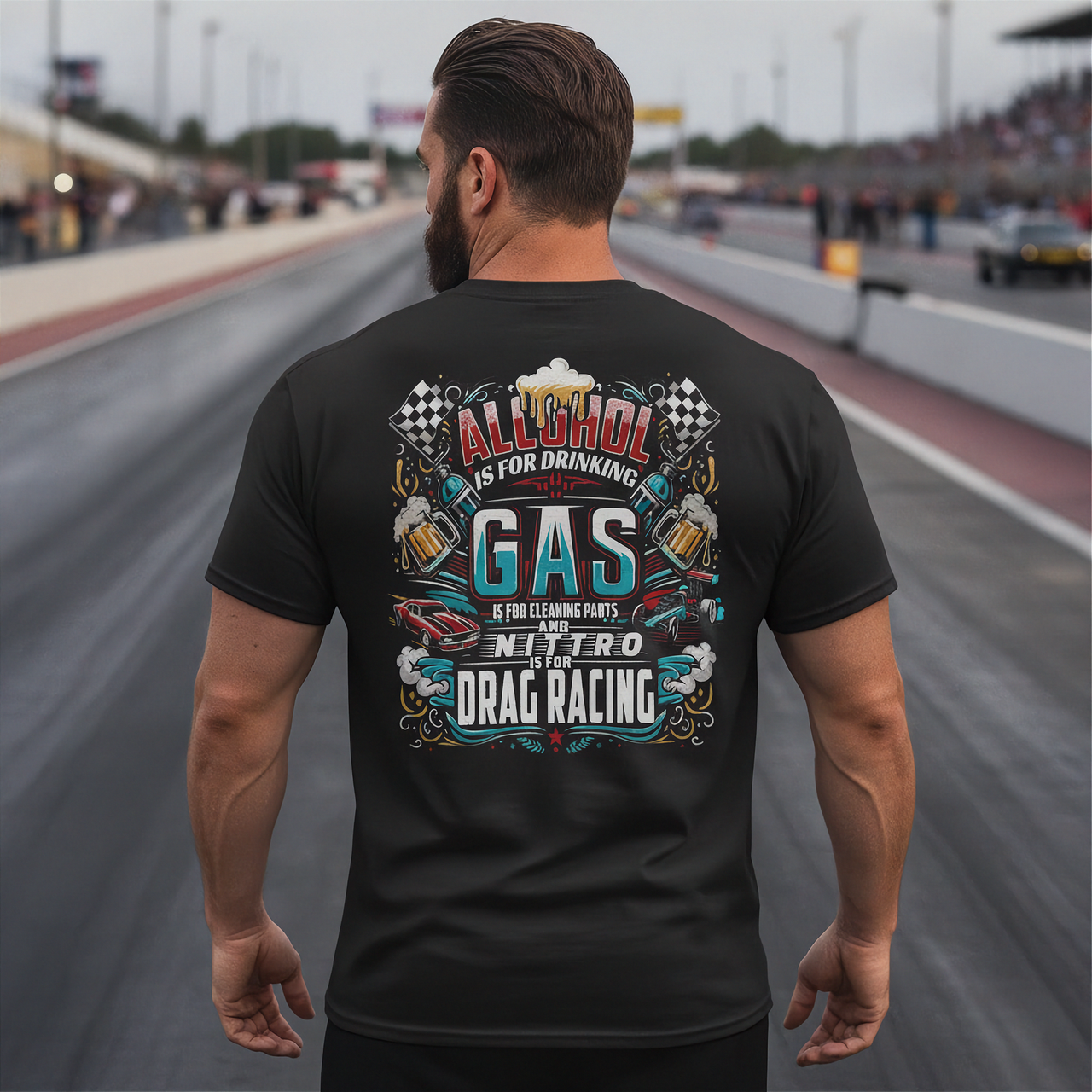 A man stands on a racetrack from behind, wearing a black t-shirt with a large colorful graphic on the back. The graphic features the text 'ALCOHOL IS FOR DRINKING, NITRO IS DRAG RACING' with illustrations of beer mugs, checkered flags, and a race car.