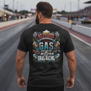A man stands on a racetrack from behind, wearing a black t-shirt with a large colorful graphic on the back. The graphic features the text 'ALCOHOL IS FOR DRINKING, NITRO IS DRAG RACING' with illustrations of beer mugs, checkered flags, and a race car.
