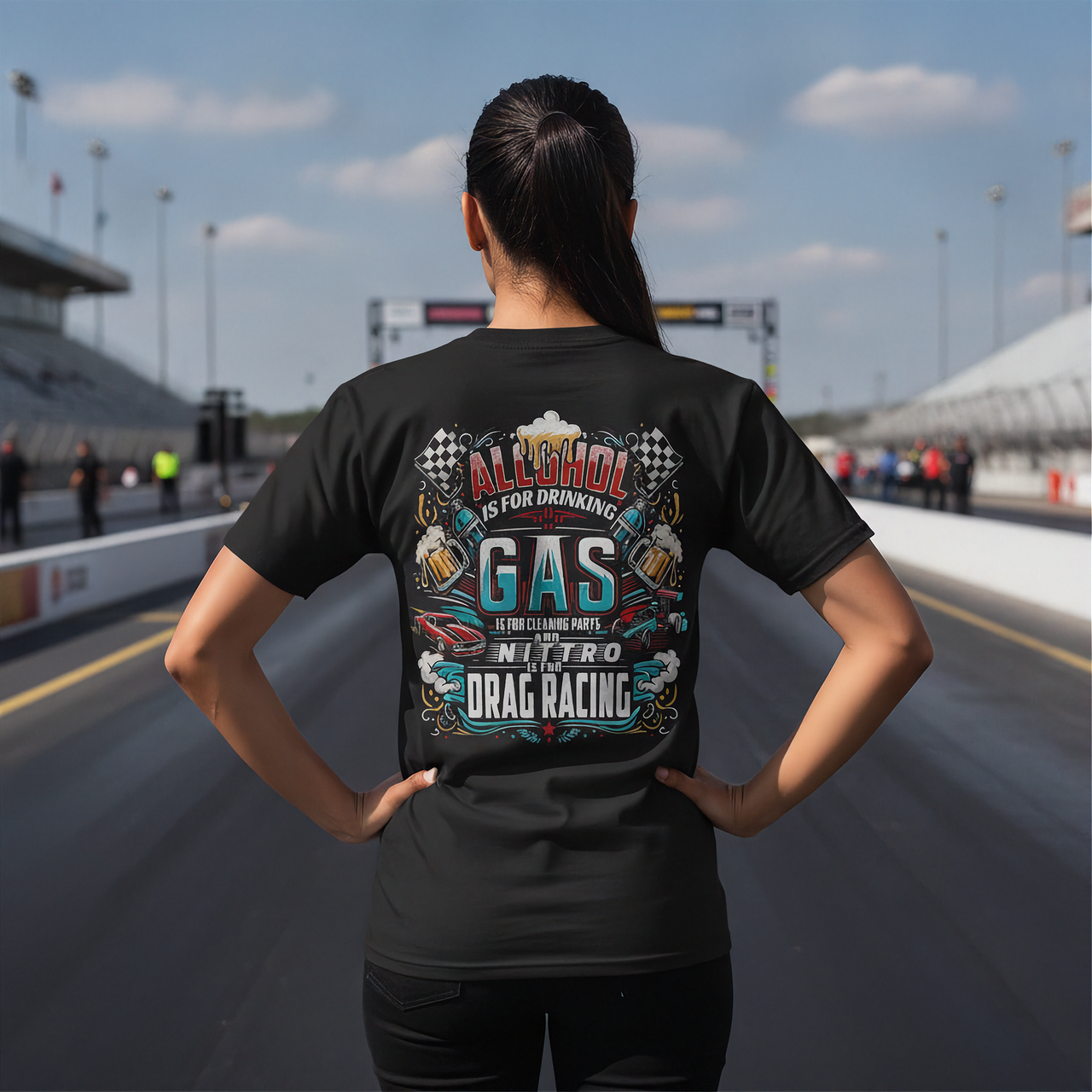 A woman stands on a racetrack from behind, wearing a black t-shirt with a colorful graphic on the back that reads 'ALCOHOL IS FOR DRINKING, NITRO IS DRAG RACING'.