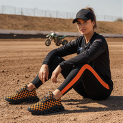 Woman sitting on a dirt track wearing black and orange shoes, black outfit with orange stripe, and cap.