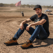 Man sitting on a dirt track wearing checkered shoes, black t-shirt, and cap.