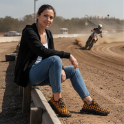 Woman sitting on a barrier at a dirt track wearing orange checkered shoes with a rider in the background
