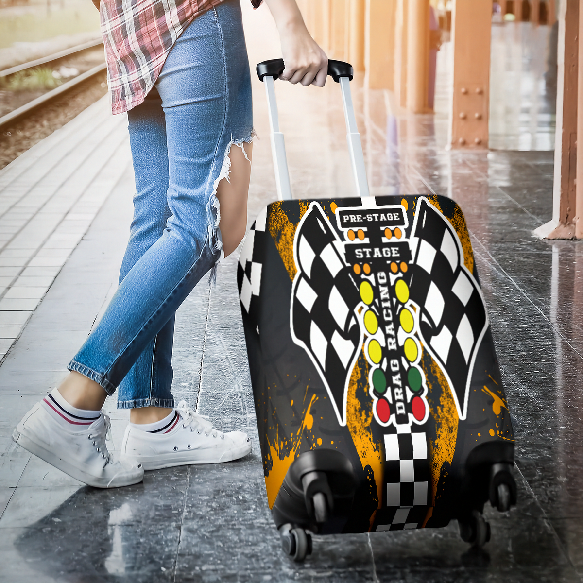 A person in a plaid shirt and ripped blue jeans is pulling a rolling suitcase on a train platform. The suitcase has a printed cover featuring a black-and-white checkered flag design, a racing light bar with yellow, green, and red lights, and orange paint-splatter accents.