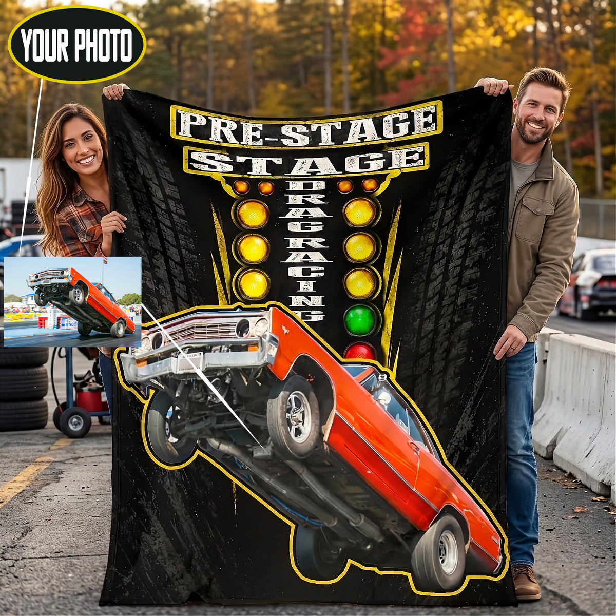 A man and a woman outdoors hold up a large black custom blanket or poster featuring a red muscle car doing a front flip, with the text 'PRE-STAGE DRAG RACING' and illuminated race lights. The photo is composited over a street scene with a small inset showing a car lift setup.