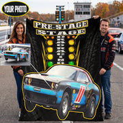 A custom drag racing themed blanket held up by a man and a woman on an outdoor racetrack. The blanket features a black background with a tire tread pattern, a yellow and black 'PRE-STAGE DRAG RACING' sign, and a blue, black, and red Dodge Challenger race car graphic.