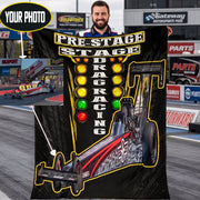 A man stands on a racetrack holding a large custom drag racing graphic print or poster. The print features a black background with a stylized drag racer car, illuminated light bar, and the words 'PRE-STAGE DRAG RACING.