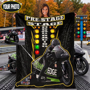 A woman holds up a large black blanket or throw featuring a motorcycle racer on a track, with the words 'PRE-STAGE DRAG RACING' and a yellow-green checkered light graphic. The background shows a racetrack scene with motorcycles and a traffic light, plus a small inset photo of a woman's photo frame.