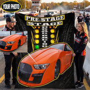 A custom printed drag racing themed blanket or poster being held up by two people on a racetrack. The design features a large orange Shelby Mustang car, a yellow and black pre-stage lighting graphic, and the words 'PRE-STAGE DRAG RACING'.