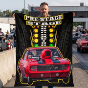 A man stands outdoors holding up a large black printed blanket or poster featuring a red muscle car, yellow stage lights, and the words 'PRE-STAGE DRAG RACING'. The background shows a real outdoor drag racing environment with cars and a concrete barrier.
