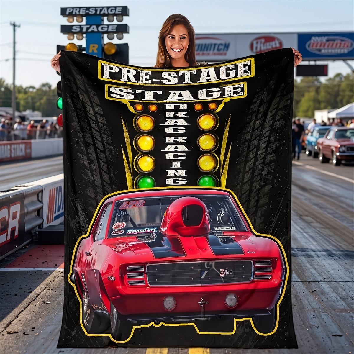 A woman stands on a drag racing track holding a large black printed blanket or poster featuring the words 'PRE-STAGE DRAG RACING' above a red race car with yellow and green light graphics.
