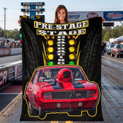 A woman stands on a drag racing track holding a large black printed blanket or poster featuring the words 'PRE-STAGE DRAG RACING' above a red race car with yellow and green light graphics.