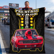 A man stands on a drag racing track holding up a large black promotional banner or poster featuring the text 'PRE-STAGE DRAG RACING' above a red muscle car graphic with yellow and green light accents. NHRA signage and racetrack buildings are visible in the background.