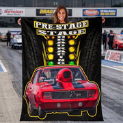A woman stands on a drag strip holding up a large black printed blanket or poster featuring a red muscle car, tire textures, and the words 'PRE-STAGE DRAG RACING.' Real racetrack scenery and other people are visible in the background, so this is Lifestyle rather than Product/Packaging.